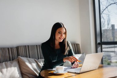 Genç ve güzel bir kız bir kafede oturuyor, bilgisayar başında çalışıyor ve sosyal ağlarda iletişim kuruyor..
