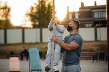 Bir baba gün batımında oğlunun maskeli bir spor sahasında ipe tırmanmasına yardım eder. Sağlıklı ebeveynlik ve sağlıklı yaşam tarzı