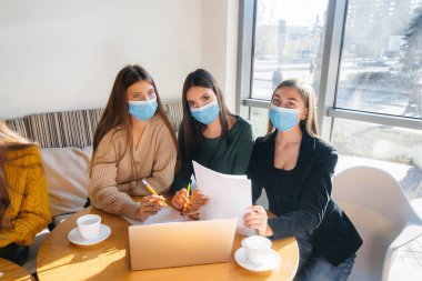 Maskeli bir grup kız bir kafede oturup dizüstü bilgisayarlarla çalışıyor. Öğrenciler.