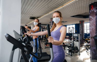 Salgın sırasında maske takan iki güzel kız spor salonunda egzersiz yapıyor. Halka açık yerlerde sosyal mesafeler.