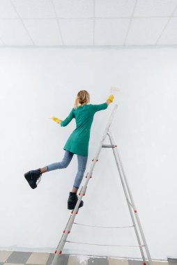 Merdivendeki genç bir kız beyaz bir duvarı silindirle boyuyor. İç kısmın tamiri.