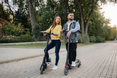 Genç ve güzel bir çift sıcak bir sonbahar gününde parkta elektrikli scooter kullanıyor. Hobiler ve eğlence..