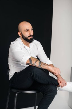 Beyaz gömlekli genç sakallı bir adam siyah arka planda oturuyor..