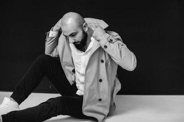 Genç sakallı bir adam siyah bir arka planda yağmurlukla yerde oturuyor. Siyah ve beyaz.