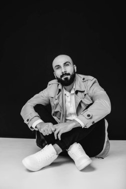 Genç sakallı bir adam siyah bir arka planda yağmurlukla yerde oturuyor. Siyah ve beyaz.