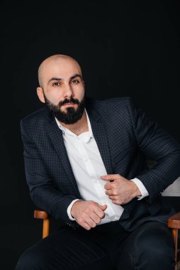 Beyaz gömlekli ve takım elbiseli genç sakallı bir adam siyah arka planda oturuyor..