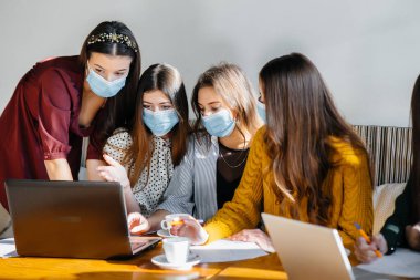 Maskeli bir grup kız bir kafede oturup dizüstü bilgisayarlarla çalışıyor. Öğrenciler.