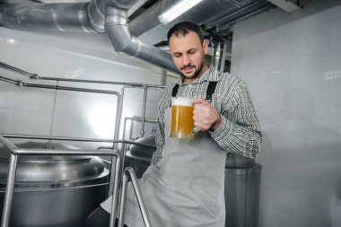 Genç sakallı bir bira imalatçısı bira fabrikasında yeni demlenmiş biranın kalite kontrolünü elinde tutuyor..
