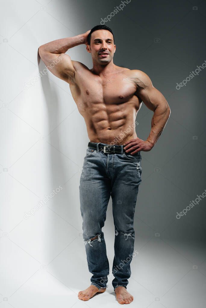 A young athlete bodybuilder poses in the studio topless in jeans near the wall. Sport.