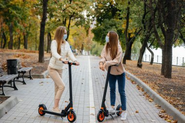 Maskeli iki genç kız sıcak bir sonbahar gününde parkta elektrikli scooterlara biniyor. Parkta yürü..
