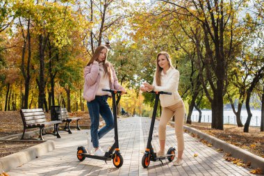 Ilık bir sonbahar gününde iki güzel kız parkta elektrikli scooterlara biniyor.