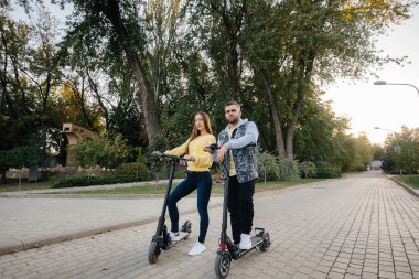 Genç ve güzel bir çift sıcak bir sonbahar gününde parkta elektrikli scooter kullanıyor. Hobiler ve eğlence..