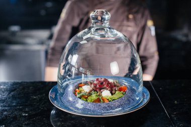 Profesyonel bir aşçı, rafine bir restoranda cam bir kubbenin altında taze domates ve soslu dana eti salatası servis eder..