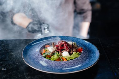 Profesyonel bir aşçı, kalın dumanlı cam kaputun altında yeni hazırlanmış domates ve dana eti salatası servis eder. Restoranda güzel dumanlı servis..