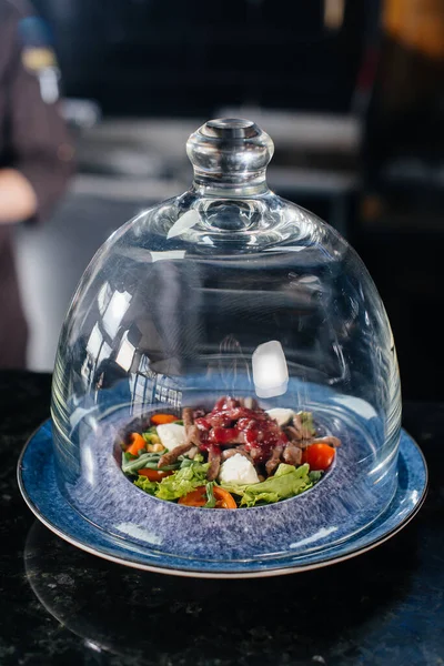 stock image A professional chef serves a freshly prepared salad of tomato and veal greens with sauce in a refined restaurant under a glass dome.