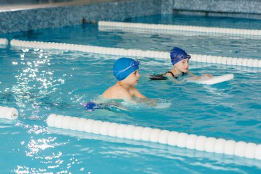 Bir grup kız ve erkek, bir eğitmenle havuzda yüzmeyi öğreniyor. Çocuk sporlarının gelişimi.