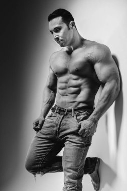 Genç bir sporcu stüdyoda üstsüz poz veriyor. Duvarın yanında kot pantolon var. Siyah ve beyaz.