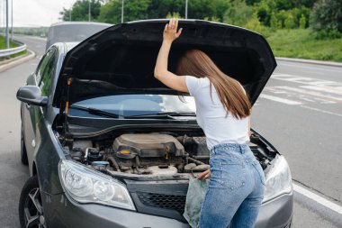 Genç bir kız otoyolun ortasında bozuk bir arabanın yanında duruyor ve kaputun altına bakıyor. Aracın bozulması ve arızası. Yardım bekliyorum..