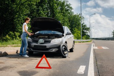 A young girl stands near a broken-down car in the middle of the highway and looks under the hood. Failure and breakdown of the car. Waiting for help. clipart