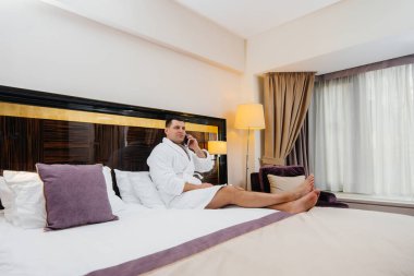 Beyaz önlüklü genç bir adam otel odasında yatağında oturmuş telefonla konuşuyor..