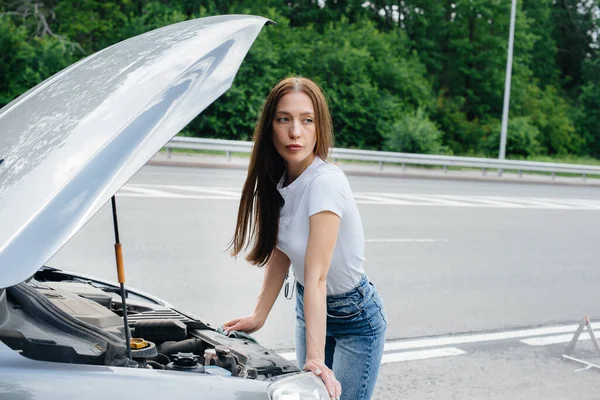 A young girl stands near a broken-down car in the middle of the highway and looks under the hood. Failure and breakdown of the car. Waiting for help.