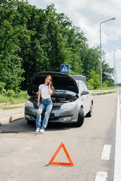 一个年轻的女孩站在高速公路中央一辆抛锚的汽车旁边 打电话求助 汽车故障和抛锚 等人帮忙 — 图库照片