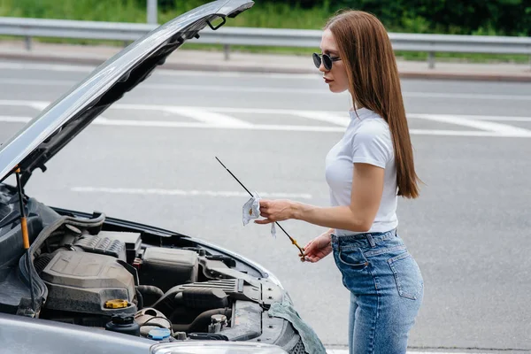 一个年轻的女孩站在高速公路中央一辆抛锚的汽车旁边 检查发动机的油耗 汽车故障和抛锚 等人帮忙 — 图库照片