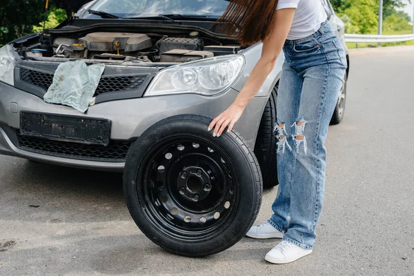 A young girl stands near a broken car in the middle of the highway and tries to change a broken wheel on a hot sunny day. Failure and breakdown of the car. Waiting for help.