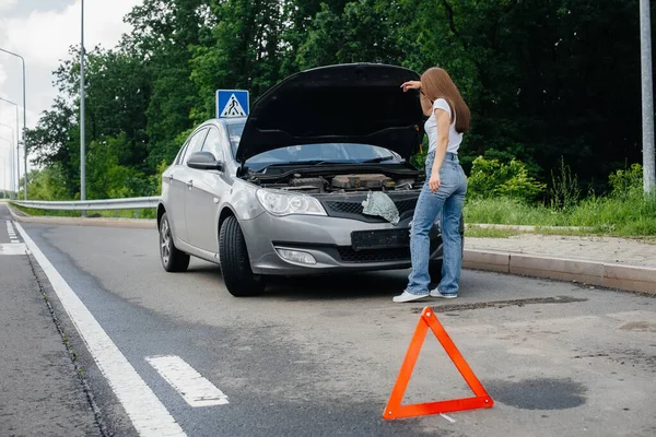 一个年轻的女孩站在高速公路中央一辆抛锚的汽车旁边 看着车篷下面 汽车故障和抛锚 等人帮忙 — 图库照片