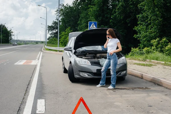 一个年轻的女孩站在高速公路中央一辆抛锚的汽车旁边 打电话求助 汽车故障和抛锚 等人帮忙 — 图库照片