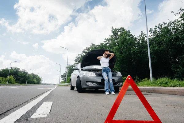 一个年轻的女孩站在高速公路中央一辆抛锚的汽车旁边 打电话求助 汽车故障和抛锚 等人帮忙 — 图库照片