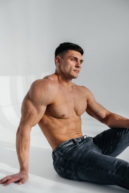 Mükemmel karın kasları olan genç seksi bir sporcu arka planda kot pantolonla stüdyoda yerde üstsüz oturuyor. Sağlıklı yaşam tarzı, düzgün beslenme, eğitim programları ve kilo vermek için beslenme.