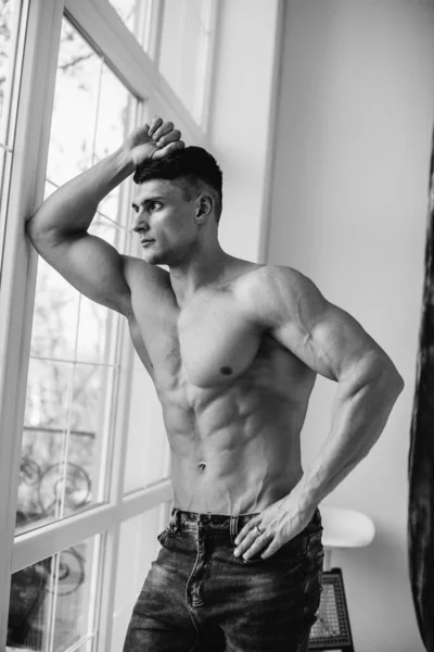 A young sexy athlete with perfect abs poses near the window in the studio topless in jeans. Healthy lifestyle, proper nutrition, training programs and nutrition for weight loss. Black and white.