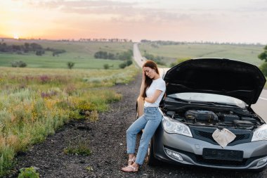 A frustrated young girl stands near a broken-down car in the middle of the highway during sunset. Breakdown and repair of the car. Waiting for help. clipart