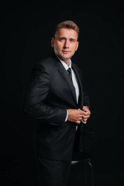 Yakışıklı bir genç adam şık bir siyah takım elbise giyer ve siyah arka planda gülümser. Temizlen, hazırlan..