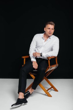 Beyaz gömlekli şık bir genç adam siyah arka planda yönetmen koltuğunda oturuyor. Yakışıklı, karizmatik bir iş adamı..