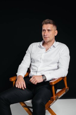 Beyaz gömlekli şık bir genç adam siyah arka planda yönetmen koltuğunda oturuyor. Yakışıklı, karizmatik bir iş adamı..