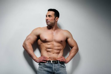 Genç bir sporcu stüdyoda üstsüz poz veriyor. Duvarın yanında kot pantolon var. Spor..