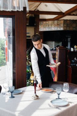 Şık üniformalı genç bir erkek garson güzel bir gurme restoranında masaya servis yapmakla meşgul. Restoran faaliyetleri, en üst düzeyde..