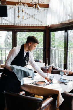 Şık üniformalı genç bir erkek garson güzel bir gurme restoranında masaya servis yapmakla meşgul. Restoran faaliyetleri, en üst düzeyde..