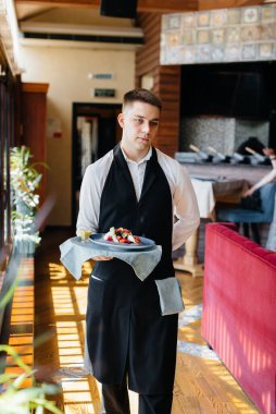 Şık bir üniforma içindeki genç bir garson, güzel bir restoranın yakın çekiminde masanın yanındaki tepside nefis bir tabakla duruyor. Restoran faaliyetleri, en üst düzeyde..