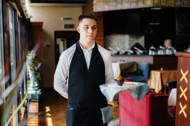 Şık bir üniforma içindeki genç bir garson, güzel bir restoranın yakın çekiminde masanın yanındaki tepside nefis bir tabakla duruyor. Restoran faaliyetleri, en üst düzeyde..