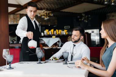 Zarif önlüklü genç bir garson zarif bir restoranda güzel bir çiftle masa servisi yapıyor. Müşteri hizmetleri.