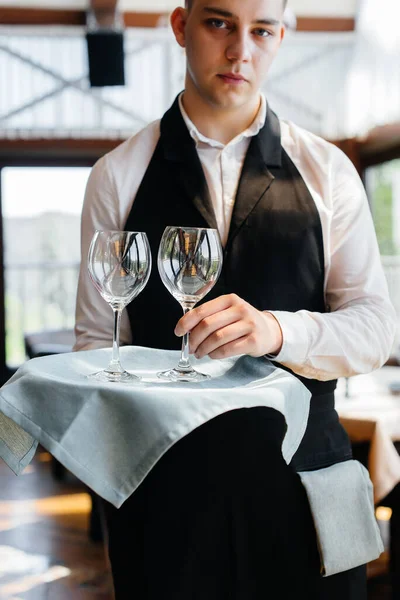 スタイリッシュな制服を着た若いウエイターが テーブルの近くのトレイに眼鏡をかけ 美しいグルメレストランの近くに立っています 最高レベルのレストラン活動 — ストック写真