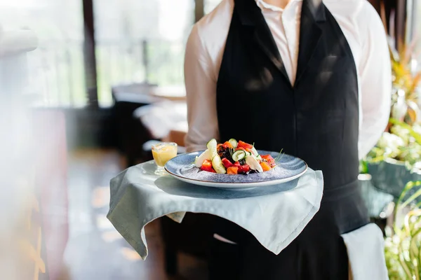 一位穿着时髦制服的年轻侍者站在一家漂亮餐厅的近旁 盘子里有一道精美的菜 最高层次的餐厅活动 — 图库照片