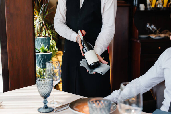 A young waiter in a stylish apron demonstrates and offers a fine wine to a client in a restaurant. Customer service.