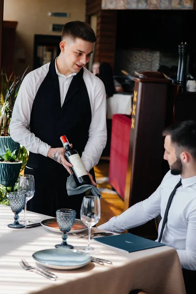 スタイリッシュなエプロンの若いウエイターは レストランの顧客に素晴らしいワインを実証し 提供しています カスタマーサービス — ストック写真