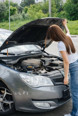 Genç bir kız otoyolun ortasında bozuk bir arabanın yanında duruyor ve kaputun altına bakıyor. Aracın bozulması ve arızası. Yardım bekliyorum..