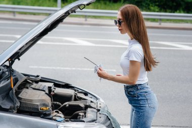 Genç bir kız otoyolun ortasında bozuk bir arabanın yanında duruyor ve motorun yağ seviyesini kontrol ediyor. Aracın bozulması ve arızası. Yardım bekliyorum..