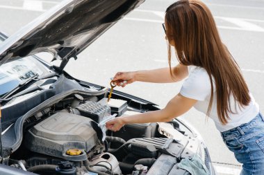 A young girl stands near a broken-down car in the middle of the highway and checks the oil level in the engine. Failure and breakdown of the car. Waiting for help. clipart